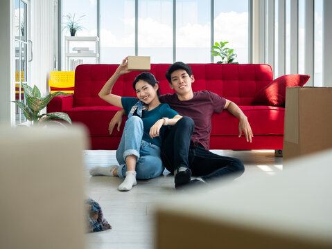 Asian Young Couple Boy And Girl Relaxed Sitting After Break From Carrying And Opening Cardboard And Parcel Box In New Home Together While She Holding Relocation Gift From Neighbor In Living Room