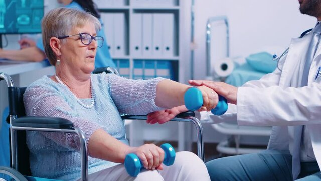 Physiotherapy for elderly woman in wheelchair, she is practicing with dumbbells to recover after a musle trauma. Modern private hospital or rehabilitation clinic. Lifting exercise for disabled people