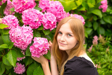 Fototapeta premium Beautiful young blonde woman with flowers near face. Pink Hydrangea macrophylla blooming in summer in botanical garden