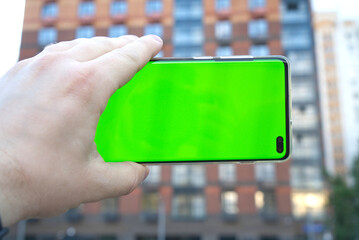 a man's hand holds a phone with a green screen chroma key on the background of a blurred building