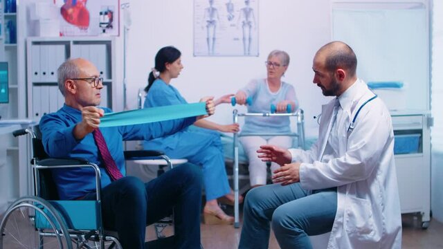 Elderly disabled man in wheelchair training muscle strength with professional doctor in modern recovery clinic or hospital. Invalid physiotherapy program, health care injury rehabilitation
