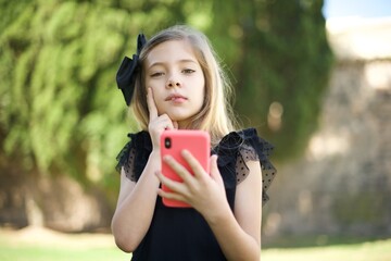 Image of a thinking dreaming young beautiful  little girl  posing isolated over pink wall background using mobile phone.
