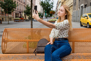 Beautiful young woman with blonde hair taking selfie at the city street background. Girl sitting on a bench in a sunny day. Active life concept