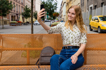 Beautiful young woman with blonde hair taking selfie at the city street background. Girl sitting on a bench in a sunny day. Active life concept