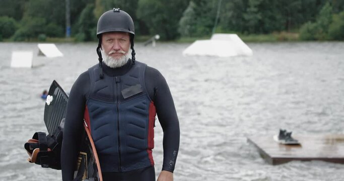 Wakeboarder, portrait of adult man, sportsman stands near the lake, looks at the camera and holds a wakeboard in his hands, active lifestyle and water sports.