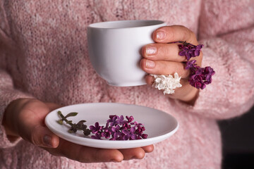 Woman's hand holding a white cup of coffee. With a beautiful lilac flowers. Drink, fashion, morning