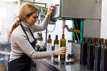 Woman working on bottling conveyor of food producing factory, filling glass bottles with olive oil