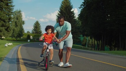 Caring affectionate handsome african american father teaching cute preschooler mixed race son with curly hair to ride bicycle in public park. Little child with help of dad learning cycling outdoors.