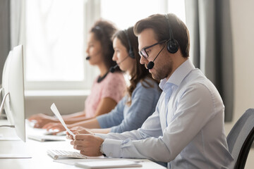 Serious call center agent in wireless headset holding, reading paper documents, sitting at...