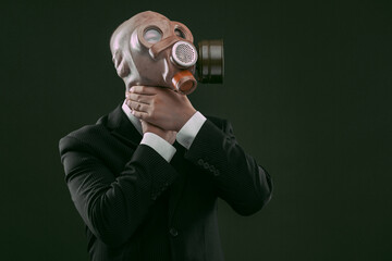 Portrait of a man wearing a dark business suit and a gas mask while choking himself, isolated on dark green background.