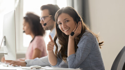 Smiling woman call center operator in headset showing thumbs up gesture, sitting at workplace in customer support service office, woman in headphones looking at camera, horizontal photo