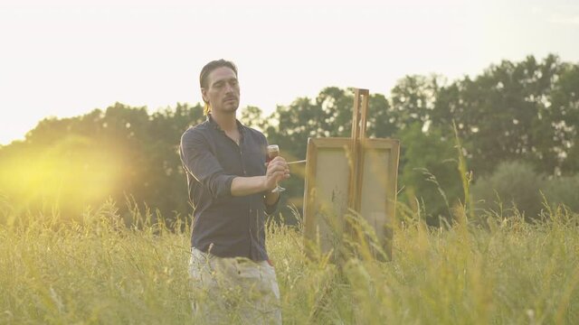 Concentrated Caucasian man painting landscape at sunset. Portrait of inspired male painter with wineglass drawing picture outdoors on summer day. Process of artwork creation.