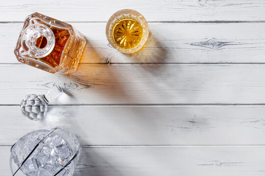 View Of A Crystal Glass And Decanter Full Of Golden Whisky, And Crystal Ice Bucket, Shot From Above On A Distressed White Wooden Background With Copy Space