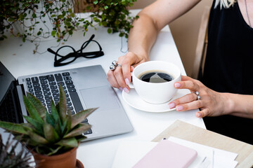 Woman's hand holding cup of coffee in cozy summer office with laptop and green plants