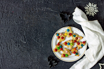 Christmas cookies of a Christmas tree with multicolored dragees on a white plate on a dark background, next to a napkin, baking pots, flat lay, copy space, top view.  Christmas baking cookie concept