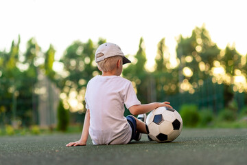 Naklejka premium a boy in sports clothes sits on a green lawn on a football field with a soccer ball on his back at sunset, rear view, sports section, training