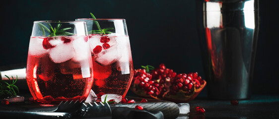 Red cocktail with cold vodka and pomegranate juice, ice cubes and rosemary, bar tools, blue black...