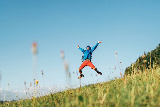 Backpacker traveler emotionally jumping over green grass mountain meadow with backpack with wide opened arms and legs. Human's freedom in nature concept image.