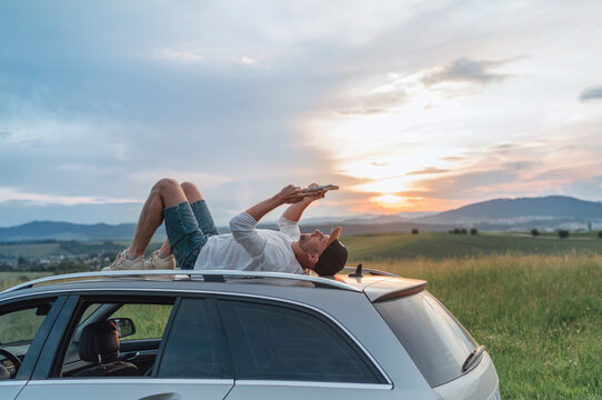 Young Man lying on the car roof and reading the paper bestseller book.He stopped his auto on the meadow with a beautiful valley view before the sunset. Reading as a hobby or education concept image