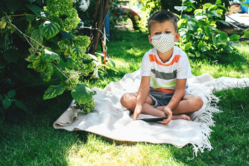 Little boy in a mask, sitting on a blanket and holds phone in hands. Quarantine, new normal concept