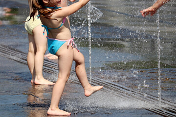 Children playing with water jets of fountain in a park. Little girl enjoying the summer, heat in city