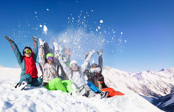 Group of young adults throw snow up in the air sitting on top of the mountain over sunny alpine peaks