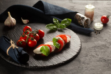 Caprese salad on stone plate with Mozzarella,fresh basil leaves,tomatoes,sea salt,dried pepper and fresh garlic on dark background.