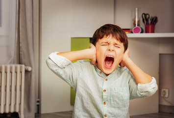 Little autistic boy with strong negative face expression closing ears and loudly screams - obrazy, fototapety, plakaty