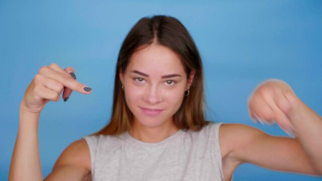 Positive woman in gray t-shirt smiles, shows middle finger on blue background with copy space. 4K slow motion footage