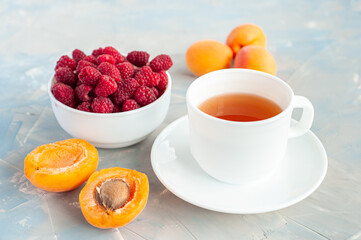 Summer light diet breakfast. A cup of green tea with ripe berries and fruits. Raspberries in a bowl and orange apricots on a concrete gray-blue background
