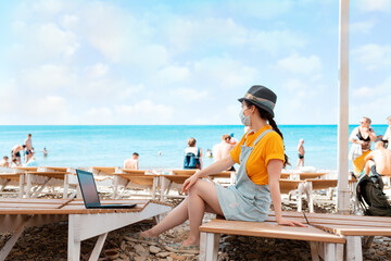 A young woman is sitting on a chaise longue in a medical mask, next to her laptop. Beach and sea in...