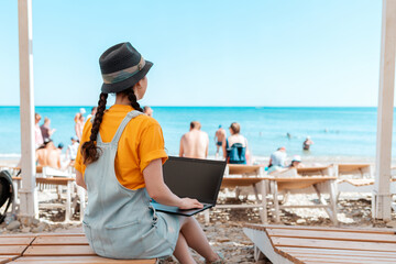 A young woman is sitting on a chaise longue and holding a laptop. Beach and sea in the background....