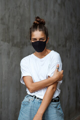 A beautiful woman in a black medical self-defense mask stands against the background of a gray concrete wall. Emotional woman looks defiantly.