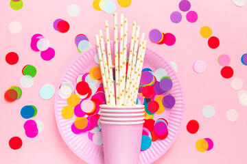 Birthday or party paper glasses with straws, dish and colorful confetti on pink background.