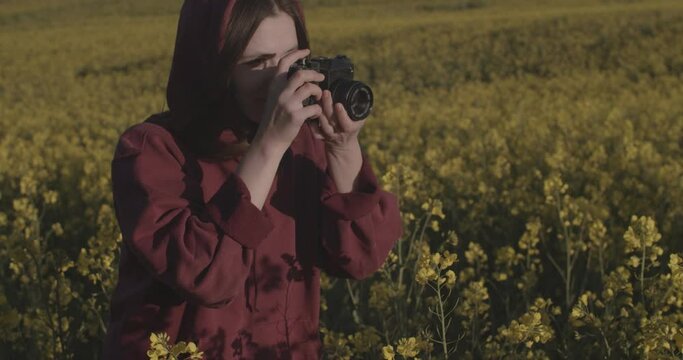Happy female photographer focusing on vintage camera outdoors on shallow nature background. Caucasian girl shooting alone making photos of yellow flower field. Hobby photographing equipment