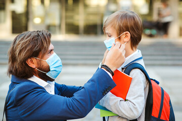 Dad putting on a protective mask on his son's face outdoors. Schoolboy with a backpack. Back to...