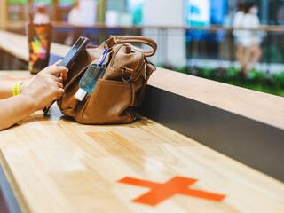 Mini alcohol gel bottle to kill Corona Virus(Covid-19) hang on a leather shoulder bag of a woman wearing a mask while use smartphone with alternative seating mark for social distancing in the mall.