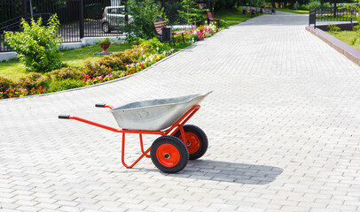 Side view on a metallic wheelbarrow with red wheels standing on paved path in blooming public park