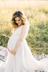 Fototapeta na wymiar Pretty caucasian pregnant woman in white dress sitting on a picnic blanket on a meadow at sunset. Summer evening in nature. Happy maternity and pregnancy concept