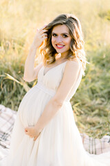 Fototapeta na wymiar Pretty caucasian pregnant woman in white dress sitting on a picnic blanket on a meadow at sunset. Summer evening in nature. Happy maternity and pregnancy concept