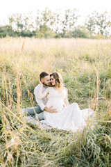 Pretty young pregnant woman with her handsome bearded man resting on a blanket in the summer meadow. Happy pregnant couple sitting on a blanket in a field