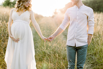 Fototapeta na wymiar Young happy romantic pregnant couple walking in wild field in summer day. Pregnant woman in white dress and her handsome man expecting a baby, enjoying the walk in summer field