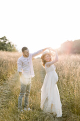 Fototapeta premium Young Caucasian couple in love walking and dancing in beautiful summer field. Pregnant woman in hat and white dress dansing with her handsome man. Happy moments. and pregnancy concept.