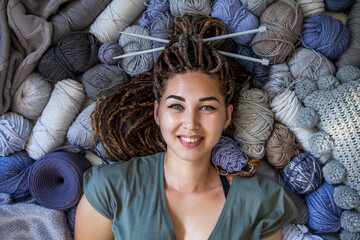 A beautiful young girl knitter lies and smiles surrounded by yarn for knitting. Female hobby and handicraft concept. Woman with dreadlocks.