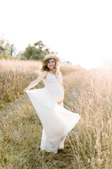 Fototapeta na wymiar Pretty pregnant girl in white long dress and in straw hat walking in the summer field on a sunset. Beautiful pregnant woman in the summer nature meadow