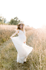 Pretty pregnant girl in white long dress and in straw hat walking in the summer field on a sunset. Beautiful pregnant woman in the summer nature meadow