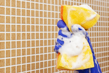 Hand in blue glove compresses the yellow sponge and soap in foam on a background of brown cells
