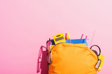 Yellow backpack packed with school stationery and eyeglasses on pink