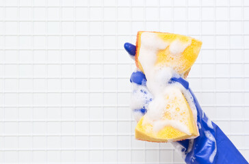 Hand in blue glove compresses the yellow sponge in the foam on a background of white tiles