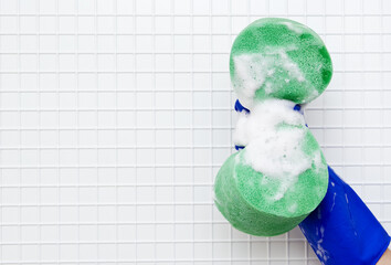 Hand in blue glove compresses green sponge in the foam on a background of white tiles